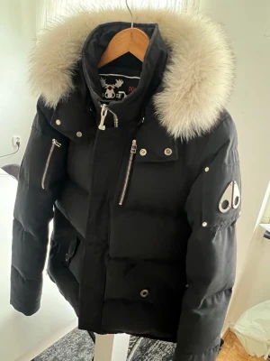 Mörk blå/svart Moose Knuckles jacka  - Snygg svart dunjacka från Moose Knuckles med stor fluffig pälskant på huvan, silverfärgade dragkedjor och knappar samt logga på ärmen. Jackan har flera fickor och en modern, puffig passform. Perfekt för kalla vinterdagar.