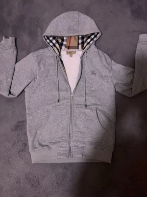 Grå zip hoodie från Burberry - Snygg grå hoodie med dragkedja från Burberry. Klassisk huva med det ikoniska rutmönstret på insidan, dragsko och två stora fickor framtill. Tillverkad i mjukt bomullsmaterial.  Tags 🏷️ ingår den är helt ny. Fråga gärna om mer bilder.