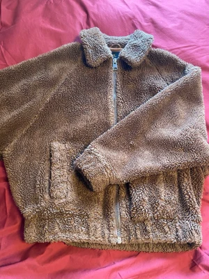 Brun teddyjacka med dragkedja - Mysig brun teddyjacka med mjukt, fluffigt material och klassisk krage. Jackan har dragkedja framtill och två stora fickor. Perfekt för kyliga dagar när du vill vara både varm och snygg.