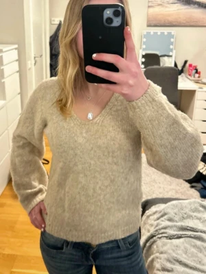 Beige stickad tröja med v-ringning - Mysig beige stickad tröja med v-ringning och långa ärmar. Tröjan har en avslappnad passform och ribbstickade muddar vid ärmslut och nederkant. Perfekt för lager på lager och enkel att matcha med jeans samt att både klä upp och klä ner.