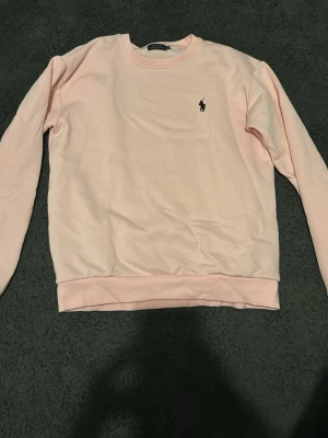 Ljusrosa sweatshirt från Polo Ralph Lauren - Ljusrosa sweatshirt från Polo Ralph Lauren med klassisk rund halsringning och broderad logga på bröstet. Tröjan har långa ärmar och ribbade muddar vid ärmslut och nederkant. Perfekt för en avslappnad och stilren look.