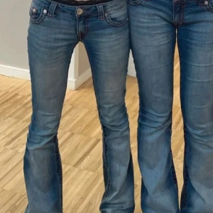 Blå bootcut jeans från True Religion - Säljer ett par klassiska blå bootcut jeans från True Religion med snygga röda sömmar och låg midja. Jeansen har fem fickor och en ikonisk logga på insidan. Perfekta för dig som gillar en tidlös och avslappnad stil.