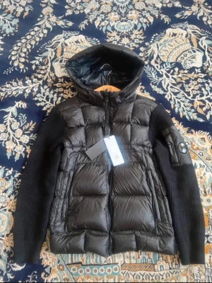 Svart dunväst med huva från CP COMPANY - Snygg svart dunväst från Cp company med stickade ärmar och vadderad kropp. Västen har en stor huva, två dragkedjefickor fram och en cp company-logga på ärmen. Materialmix med glansig dunfront och ribbstickade ärmar och rygg ger en modern look.