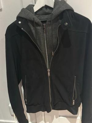 Svart bomberjacka från Zara Man - Svart bomberjacka från Zara Man med grå huva i sweatshirtmaterial. Jackan har dragkedja framtill, snörning i huvan och två fickor med dragkedja. Perfekt för en cool och avslappnad stil.