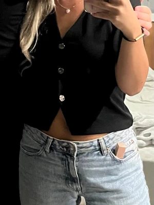 Svart kortärmad tröja med silvriga knappar - Svart tröja med v-ringning och korta ärmar. Toppen har en croppad passform och knäpps med tre knappar framtill. 