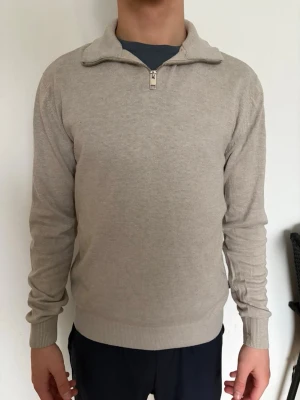 Beige stickad tröja med halvzip - Beige stickad tröja med halvzip och hög krage. Tröjan har långa ärmar och ribbade muddar vid ärmslut och nederkant. Perfekt för lager-på-lager och enkel att matcha med olika outfits.