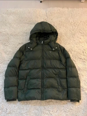 Mörkgrön pufferjacka från Gant  - Snygg mörkgrön pufferjacka från Gant med avtagbar huva och dragkedja framtill. Storlek S. Fint skick.