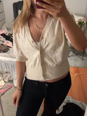 Beige kortärmad kofta med mönster - Snygg beige kofta med korta ärmar och diskret mönster. Koftan har v-ringning, knäppning framtill och en croppad passform. Perfekt att styla över ett linne eller t-shirt för en trendig look.