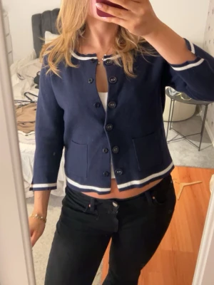 Mörkblå kofta med vita detaljer - Snygg mörkblå kofta med vita ränder vid ärmslut, hals och nederkant. Koftan har rund halsringning, knappar framtill och två fickor. Perfekt för en stilren och klassisk look.