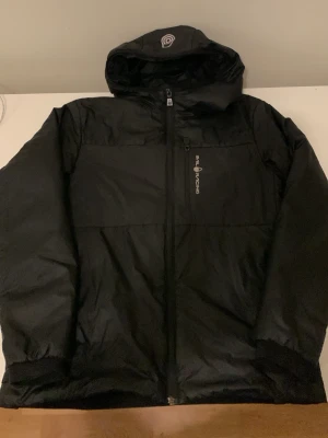 Svart pufferjacka från SEA RACING - Snygg svart pufferjacka från SEA RACING med huva och dragkedja framtill. Jackan har en diskret logga på bröstet och är tillverkad i ett glansigt, vadderat material som håller värmen under kalla dagar. Perfekt för dig som vill ha en stilren och funktionell vinterjacka.