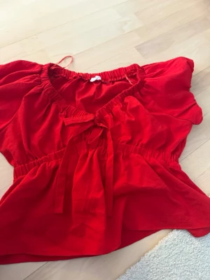 Röd blus med knyt från Enna Sky - Snygg röd blus från Enna Sky med korta puffärmar och elastisk midja. Toppen har en söt knytning framtill och en luftig passform som ger en avslappnad men trendig look. Perfekt att styla med jeans eller kjol för en färgstark outfit.