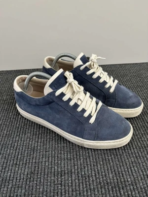  Brunello Cucinelli mockasneakers - Snygga sneakers i blå mocka från Brunello Cucinelli med vita detaljer och vita snören. Klassisk låg modell med vit sula och diskret logga på plösen. Perfekta för dig som gillar stilrena och exklusiva skor i premiumkvalitet.