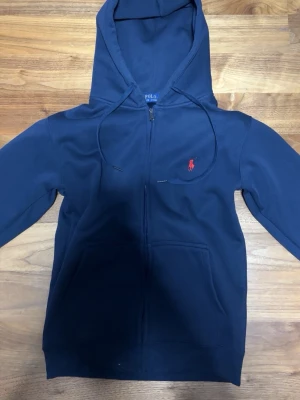 Mörkblå hoodie från Polo Ralph Lauren - Storlek S fast känns som en XS speciellt runt magenMörkblå hoodie med dragkedja från Polo Ralph Lauren. Klassisk design med huva och snören, samt fickor framtill. Materialet är mjukt och bekvämt, perfekt för chill dagar. Ikonisk röd logga broderad på bröstet ger en snygg touch. 