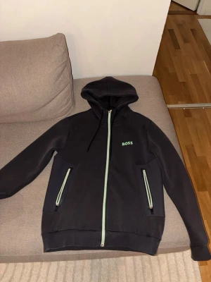 Svart hoodie med dragkedja från BOSS - Svart hoodie från BOSS med dragkedja framtill och två fickor med ljusgröna detaljer. Klassisk huva med snörning och BOSS-logga på bröstet. Perfekt för en avslappnad och stilren look. PRIS GÅR Å DISKUTERA VID SNABB AFFÄR 