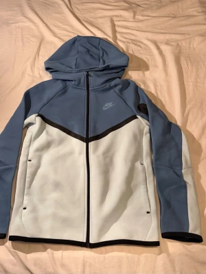 Nike blå och vit hoodie storlek 147-158 - Nike hoodie i blått och vitt med hel dragkedja framtill och svart kantband. Hoodien har en klassisk huva, två sidofickor och Nike-logga på bröstet. Materialet är mjukt och stretchigt, perfekt för chill eller träning.