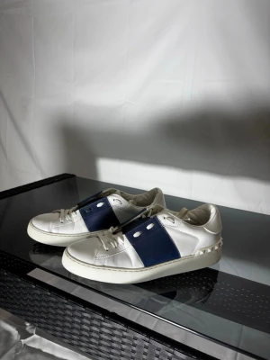 Vit och blå sneaker från Valentino - Sick 8/10.                                                                 Säljer ett par snygga sneakers från Valentino med vit läderbas och en bred mörkblå panel på sidan. Skorna har klassiska snörningar och coola nitar längs hälen. Perfekt för dig som vill ha en stilren och trendig look.