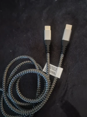 Flätad USB-C till USB-C kabel - Säljer en grå och svart flätad USB-C till USB-C kabel. 1 meter lång Kabeln har robust nylonhölje och silverfärgade kontakter med svarta detaljer. Perfekt för laddning och dataöverföring mellan kompatibla enheter.