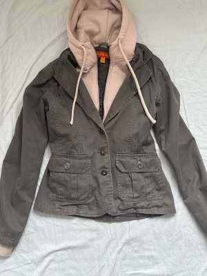 Grå kavajjacka med hoodie från Fishbone - Snygg grå kavajjacka från Fishbone med inbyggd ljusrosa hoodie och dragkedja. Jackan har två stora fickor framtill, knappar och en avslappnad passform. Perfekt för dig som vill ha en mix mellan street och casual stil.