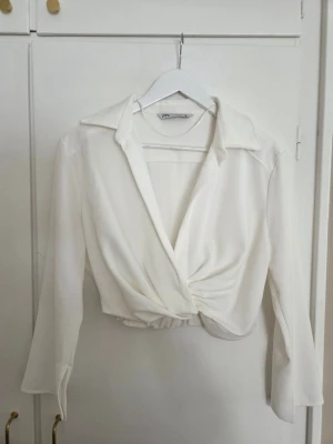 Vit omlottblus från Zara - Vit omlottblus från Zara med krage och långa ärmar. Blusen har en croppad passform och knytdetalj framtill och i sidan finns en dragkedja. 