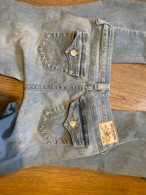 Ljusblå True Religion jeans - Säljer ett par ljusblå jeans från True Religion med ikoniska bakfickor och kontrastsömmar. Köpta secondhand men passade tyvärr inte. Jeansen har raka ben, klassisk femficksdesign och detaljerade knappar. Perfekta för dig som gillar streetwear och vill ha en avslappnad men snygg look. Små defekter, se dem sista bilderna, men inget som syns tydligt. Storlek 28💞🤗