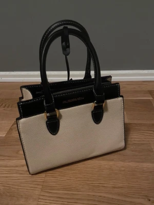Beige och svart handväska Belleborg & Mokum - Snygg beige och svart handväska från Belleborg & Mokum med guldfärgade detaljer. Väskan har två handtag, avtagbar axelrem och flera fack.