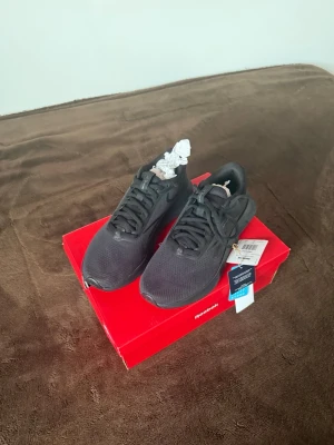 Svarta Reebok sneakers i mesh - Snygga helsvarta sneakers från Reebok med ovandel i mesh och syntetmaterial. Skorna har klassisk låg profil, svarta snören och diskret Reebok-logga på sidan. Perfekta för dig som gillar stilrena och sportiga skor.