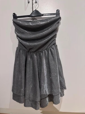 Glittrig axelbandslös klänning - Supersnygg axelbandslös klänning i glittrigt silvertyg med draperad överdel och voluminös kjol i flera lager. Perfekt för dig som vill sticka ut och glänsa på nästa event. Materialet är skimrande och ger en lyxig känsla.price tag doesn’t come with it cause its from shein but i never used it only tried and didn’t suit me