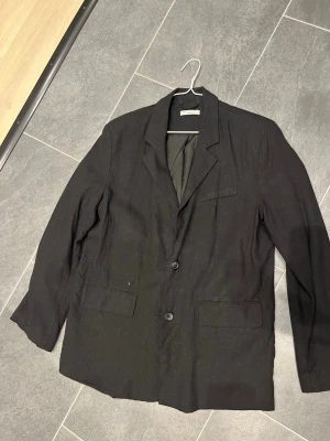 Oversized Linne Kavaj Dam Gina Tricot - Stilren svart kavaj i linne med två knappar framtill och klassisk krage. Kavajen har två fickor med lock och en bröstficka. Fodrad insida och knappar vid ärmsluten. Passar perfekt till en oversized look på sommaren.