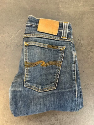 Blå jeans från Nudie Jeans - Säljer ett par klassiska blå jeans från Nudie Jeans med snygga slitningar och kontrastsömmar. Modellen har raka ben och fem fickor, samt Nudie Jeans ikoniska broderi på bakfickan. Tillverkade i mjuk denim med en skön passform.