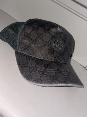 Svart keps från Gucci med GG-mönster - Snygg svart keps från Gucci med klassiskt GG-monogram över hela kepsen och metallogo framtill. Kepsen har mesh på sidorna och justerbar rem bak. Perfekt accessoar för en trendig streetwear-look.