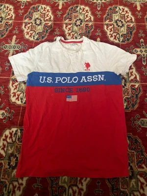 T-shirt från U.S. Polo Assn. i rött och vitt - Snygg t-shirt från U.S. Polo Assn. med vit överdel, blå band med logga och röd nederdel. Tryckt amerikansk flagga och texten 'Since 1890' framtill. Klassisk passform och tillverkad i mjuk bomull. Perfekt för en avslappnad och sportig look.