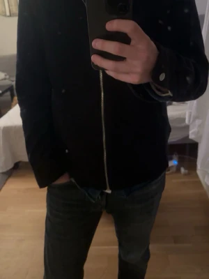 Svart bomberjacka med dragkedja - Snygg svart bomberjacka med silverfärgad dragkedja framtill och knappdetaljer vid ärmslut. Jackan har en klassisk, rak passform och är perfekt för dig som gillar en stilren look.