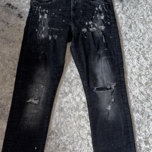 Supply jeans - Säljer ett par45 svarta jeans med slitningar, hål på ena benet och coola detaljer som färgstänk. Je passion för streetstyle och vill ha en edgy look. Klassisk femficksmodell och raka ben. Perfekta för dig som gillar en avslappnad vibe.