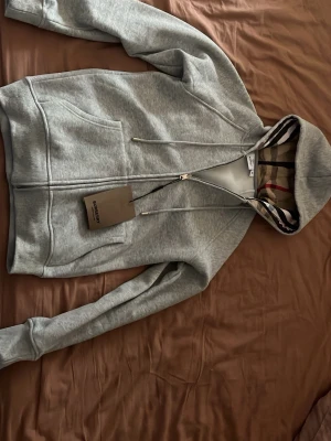 Grå Burberry zip hoodie - Snygg grå zip hoodie från Burberry med klassiskt rutigt foder i huvan och dragkedja. Tillverkad i mjukt bomullsmaterial och har två fickor framtill. Är inte helt säker på om det är en XS i kill form eller en Medium i tjej form