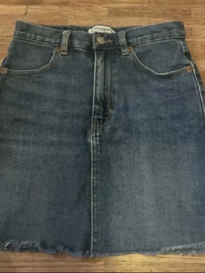 Mörkblå jeanskjol från Pull&Bear - Klassisk mörkblå jeanskjol från Pull&Bear med rå kant nertill och femficksmodell. Kjolen har bälteshällor och knappstängning framtill. Perfekt för en avslappnad och trendig look.