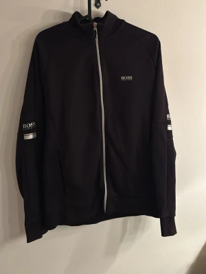 Svart zip-hoodie från BOSS - Snygg svart hoodie från BOSS med hel dragkedja och diskret logga på bröstet samt på båda ärmarna. Stilen är sportig med vita detaljer längs dragkedjan och på ärmarna. Tillverkad i mjukt material som känns bekvämt och passar perfekt till en avslappnad look.