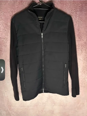 Svart quiltad cardigan från Massimo Dutti - Stilren svart cardiganjacka från Massimo Dutti med quiltad front och släta ärmar. Jackan har dragkedja framtill, två fickor med dragkedja och en hög krage. Perfekt för lager på lager och ger en clean look.