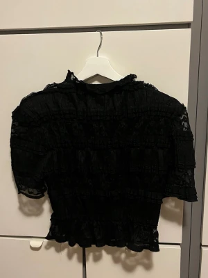 Svart spetsblus med volangdetaljer - Säljer en svart blus i spets med volangdetaljer och korta ärmar. Blusen har en romantisk look med transparenta partier och rynkade sömmar som ger extra struktur. Perfekt för dig som gillar en unik och trendig topp med mycket detaljer.