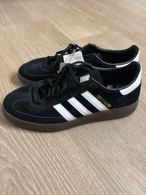 Adidas Spezial svarta sneakers - Svarta Adidas Spezial sneakers i mocka med klassiska vita ränder på sidorna och gummisula. Gulddetaljer på sidan och tungan. Lågt skaft och svart snörning. Perfekta för dig som gillar retrostil och streetwear.