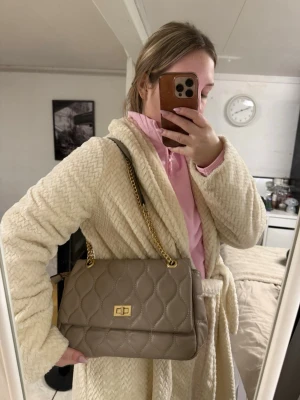Beige quiltad axelväska med kedja - Säljer en beige axelväska med quiltat mönster och guldfärgad kedja. Väskan har en rektangulär form och ett snyggt vridlås framtill. Som man ser på bilden är den smått sliten där man öppnar väskan 
