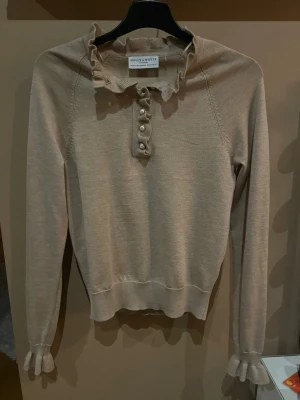 Beige stickad tröja med volang - Supersöt beige stickad tröja från Holly & White med volangdetaljer vid halsringning och ärmslut. Tröjan har pärlknappar framtill och en feminin, smal passform. Perfekt för dig som gillar romantiska detaljer och mjuka material.