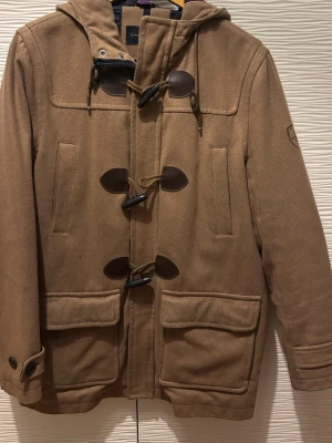 Brun duffelrock med huva från Tommy Hilfiger - Snygg brun duffelrock från Tommy Hilfiger med klassiska träknappar och läderdetaljer. Rocken har huva, två stora fickor framtill och två snedställda bröstfickor. Tillverkad i ett tjockt ullblandat material som håller värmen under kalla dagar.