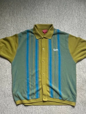 Supreme SS22 Stripe Button Up Polo - Supreme tröja i färgen grön med blå  vertikala ränder och broderad Supreme-logga på bröstet. Tröjan är kortärmad och har en retro vibe, perfekt för dig som gillar streetwear.