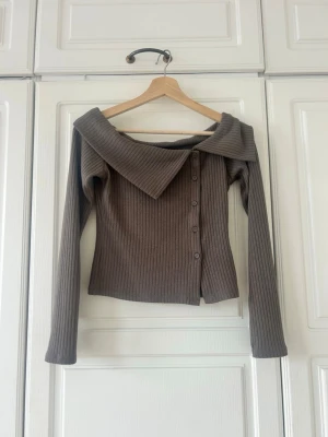 Brun ribbad offshoulder topp💖💖🥹✨ - Trendig brun ribbad topp med offshoulder-krage och dekorativa knappar längs sidan. Långärmad och figurnära passform, perfekt för dig som gillar en unik och stilren look. Materialet känns mjukt och stretchigt.