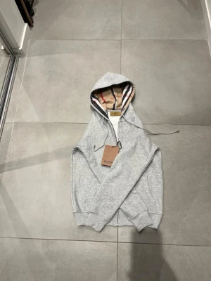 Grå zip hoodie från Burberry 100 procent cotton - Snygg grå zip hoodie från Burberry med klassiskt rutigt foder i huvan. Hoodien har dragkedja framtill, ribbade muddar och justerbar dragsko i huvan. Perfekt för dig som vill ha en stilren och exklusiv look med ikoniska Burberry-detaljer.