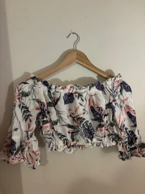 Blommig offshoulder croppad topp - Supersöt croppad offshoulder topp med blommigt mönster i vitt, lila och rosa. Toppen har långa ärmar med volangdetaljer och resår vid axlar och midja för en snygg passform. Perfekt för sommarens alla tillfällen!