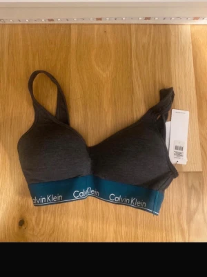 Mörkgrå bralette från Calvin Klein - Snygg mörkgrå br tagging från Calvin vith elastisk blå resår med logga. Mjuk och bekväm modell utan bygel, perfekt för dig som gillar en avslappnad stil. Justerbara axelband och lätt vadderade kupor för extra komfort.