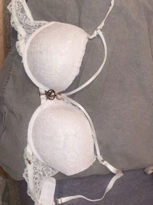 Vit push-up bh med spets och guld detaljer - Snygg vit push-up med vadderade kupor och spetsdetaljer. Bh:n har guldfärgade metallringar och ett dekorativt hänge framtill. Axelbanden är justerbara.