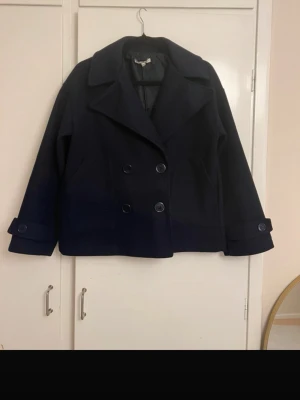 Mörkblå kappa med dubbelknäppning - Stilren mörkblå kappa med bred krage och dubbelknäppning framtill. Jackan har två sidofickor och dekorativa knappar vid ärmsluten. Klassisk rak passform och fodrad insida, perfekt för kyligare dagar.