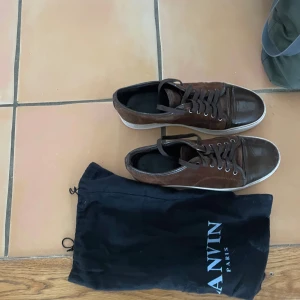 Bruna sneakers från Lanvin - Snygga bruna sneakers från Lanvin i mocka med snörning och vit sula. Perfekta för en stilren look. Skorna kommer med en praktisk dustbag för förvaring. Och så är det 7/10 i skick och är nu perfekt ny till våren allt og utom att boxen som gick sönder och ny pris  på denna skor är 5600 min MItt pris  är 1299 och kan gå ner på en snabb affär och om du är osäker på något det är bara att fråga och skon passar  bäst 44
